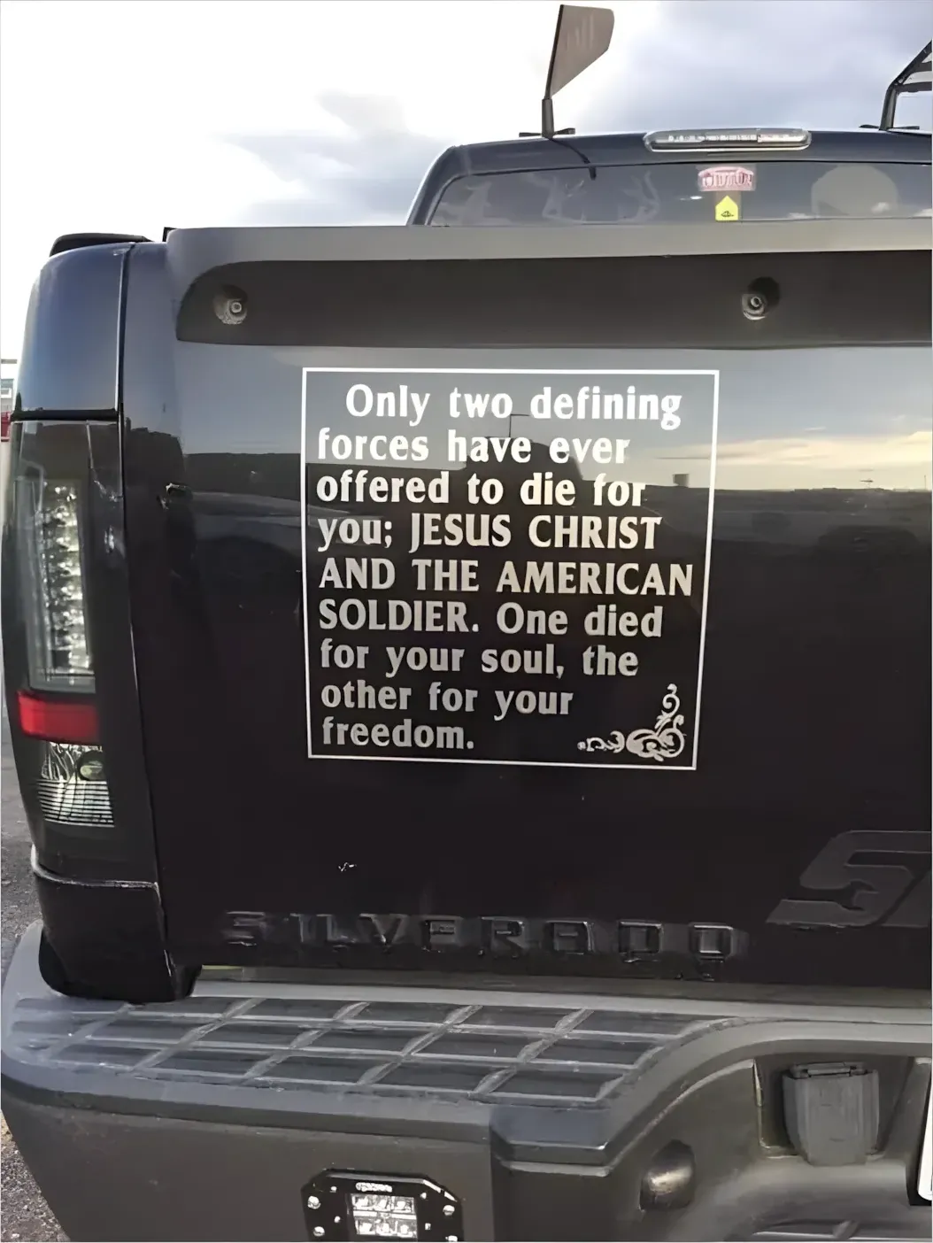 Photo of the back of a black Chevrolet Silverado truck with a large white rectangular bumper sticker reading: "Only two defining forces have ever offered to die for you: JESUS CHRIST AND THE AMERICAN SOLDIER. One died for your soul, the other for your freedom." with decorative scrollwork