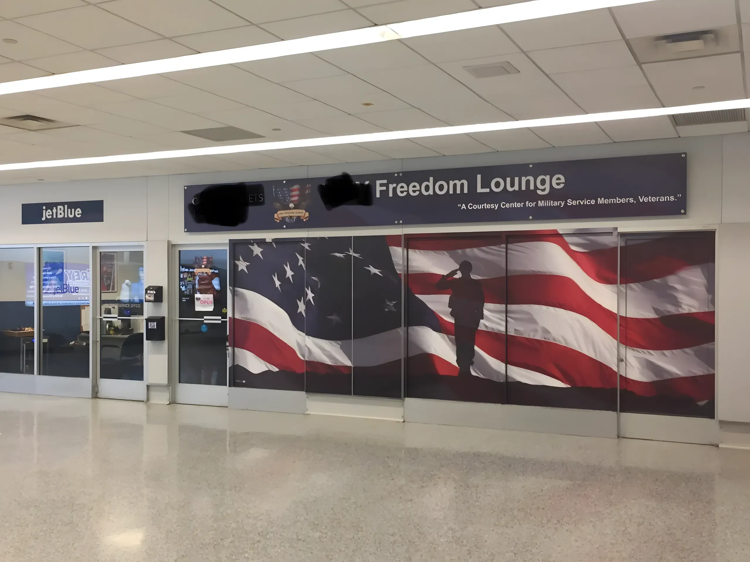 Photo of an airport terminal interior showing a "Freedom Lounge" with subtitle "A Courtesy Center for Military Service Members, Veterans." Large glass panels/doors decorated with American flag imagery and a silhouette of a saluting soldier. JetBlue counter/gate visible to the left. Fluorescent ceiling lights, polished floor. Part of the lounge name above "Freedom Lounge" is blacked out.