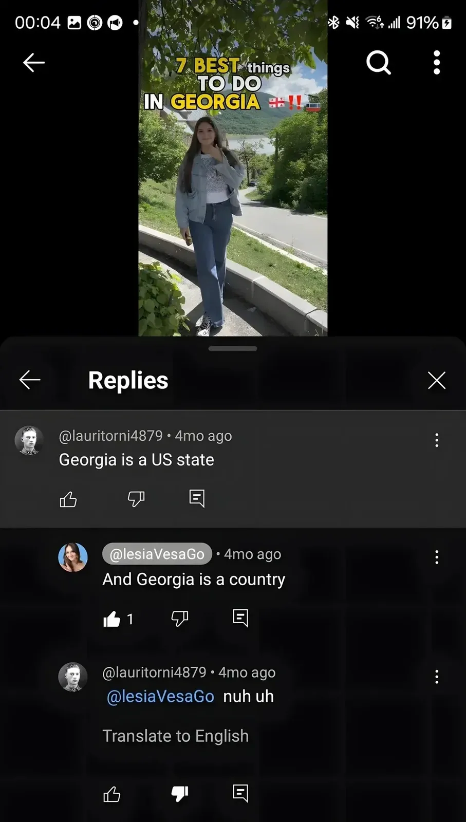 YouTube Shorts on mobile. Video thumbnail showing young woman in Georgia (the country) with scenic mountain/river backdrop and text overlay "7 BEST things TO DO IN GEORGIA" with Georgian and other flags. Replies section: @lauritorni4879 (4mo ago): "Georgia is a US state". @lesiaVesaGo (4mo ago, highlighted): "And Georgia is a country". @lauritorni4879 (4mo ago, replying to @lesiaVesaGo, downvoted): "nuh uh" (with "Translate to English" option)