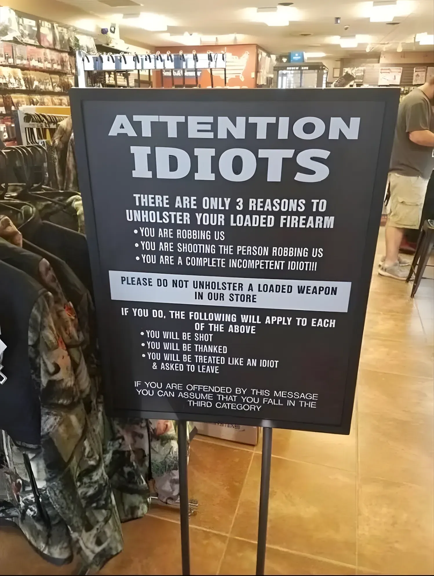 Photo of a black sign on a metal stand inside a gun/outdoor store with camouflage clothing racks visible. Sign reads: "ATTENTION IDIOTS. There are only 3 reasons to unholster your loaded firearm: You are robbing us, You are shooting the person robbing us, You are a complete incompetent idiot!!! Please do not unholster a loaded weapon in our store. If you do, the following will apply to each of the above: You will be shot, You will be thanked, You will be treated like an idiot & asked to leave. If you are offended by this message you can assume that you fall in the third category."