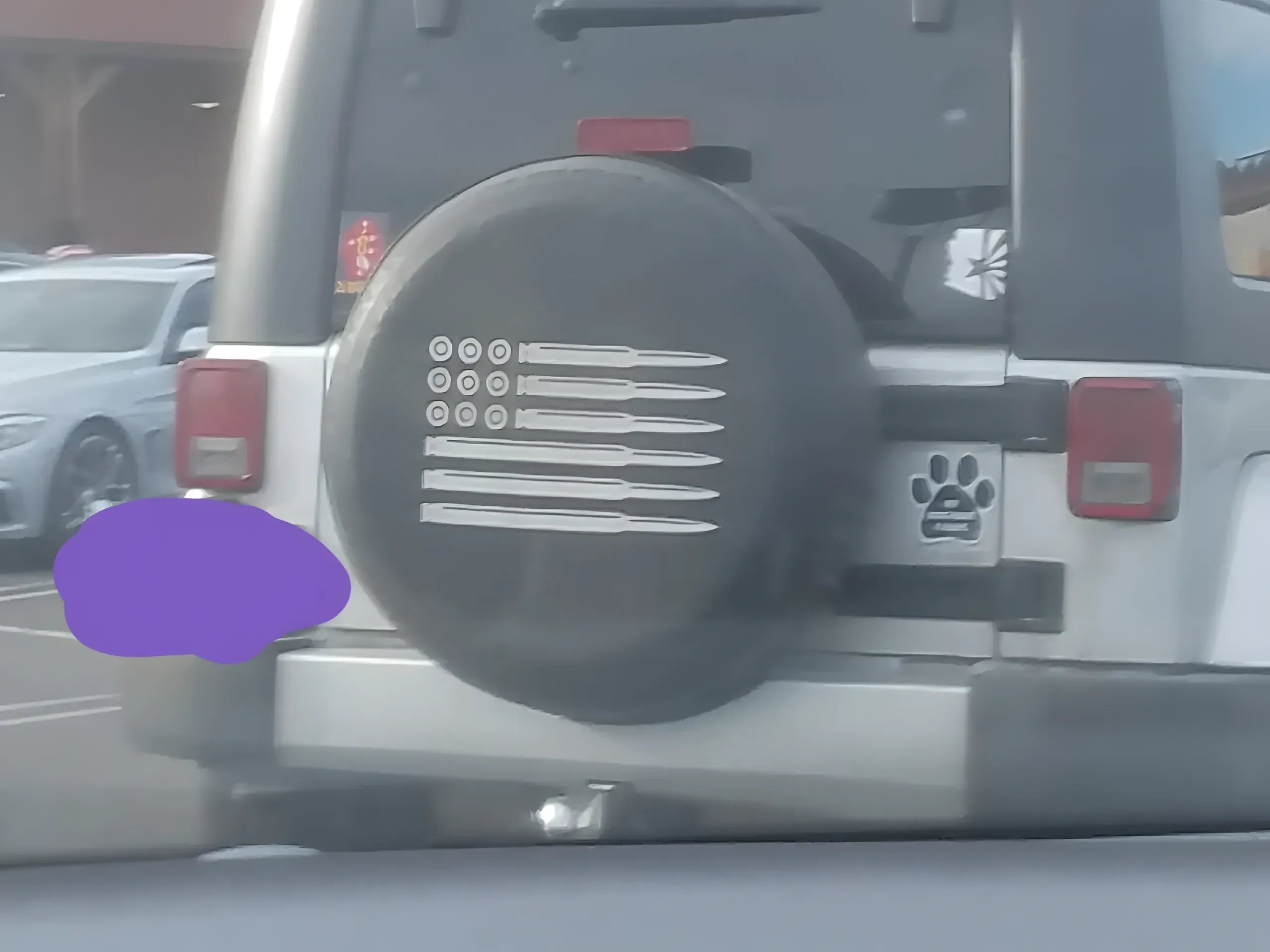 Photo taken from inside a car of the back of a Jeep Wrangler in a parking lot. The spare tire cover has an American flag design where the stars are replaced by bullet casings viewed from the end and the stripes are replaced by rifle ammunition. Purple blob covering the license plate. Paw print sticker on the vehicle body
