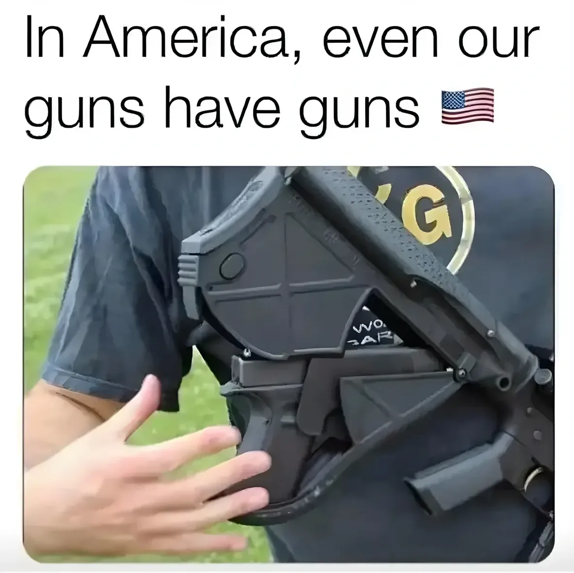 Meme. Text at top: "In America, even our guns have guns" with US flag emoji. Photo shows a close-up of a person's torso wearing a dark t-shirt, holding a large black tactical weapon that has a smaller gun mounted on it as an attachment
