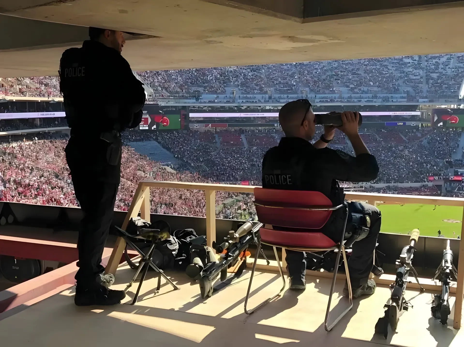 Photo of two police officers in an elevated observation/sniper position inside a sports stadium during an American football game. One officer stands looking out, another is seated looking through binoculars. Multiple rifles and equipment cases are laid out on the floor between them. Packed stadium visible through the opening