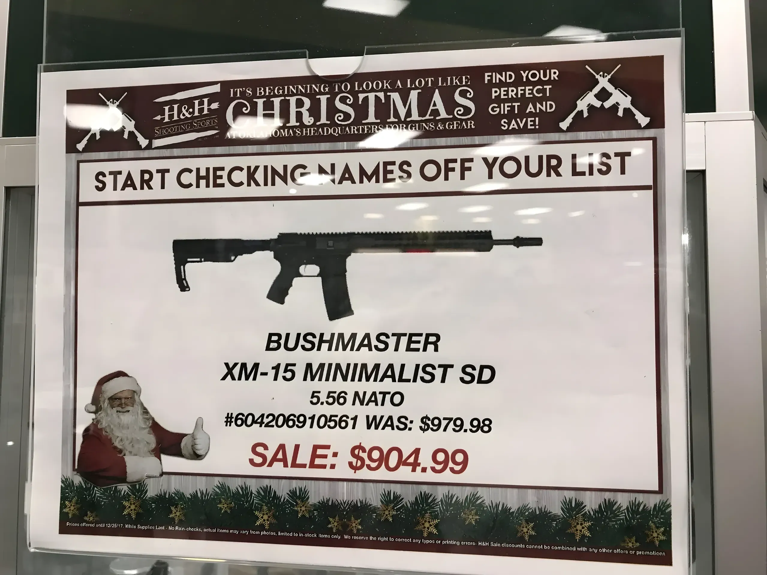 Photo of a gun store Christmas advertisement behind glass. Banner at top: "H&H - It's beginning to look a lot like Christmas - Oklahoma's headquarters for guns & gear - Find your perfect gift and save!" Below: "Start checking names off your list" with image of Santa Claus giving a thumbs up next to a rifle. Product: "Bushmaster XM-15 Minimalist SD, 5.56 NATO, #604206910561, Was: $979.98, SALE: $904.99". Holly garland decoration along the bottom