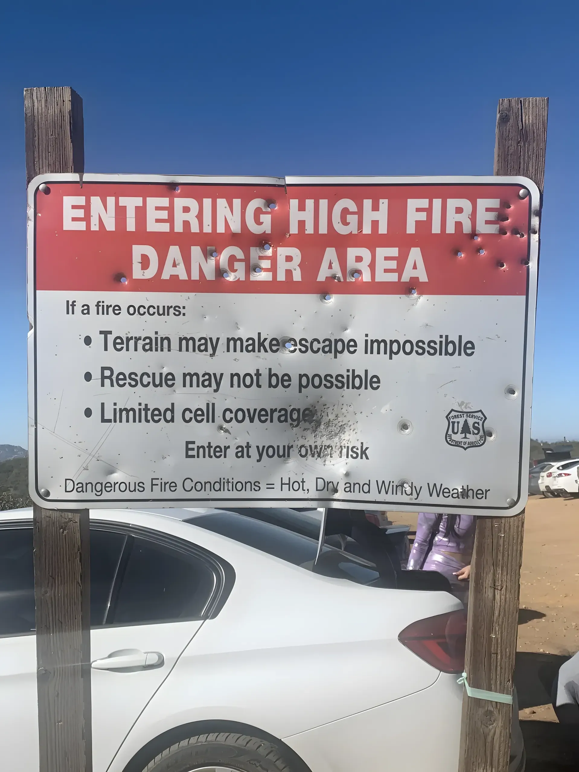 Photo of a US Forest Service sign in a rural arid outdoor area, mounted between two wooden posts. Red and white sign reads: "ENTERING HIGH FIRE DANGER AREA. If a fire occurs: Terrain may make escape impossible, Rescue may not be possible, Limited cell coverage. Enter at your own risk. Dangerous Fire Conditions = Hot, Dry and Windy Weather." USFS shield logo. The sign is riddled with bullet holes. White car visible in background