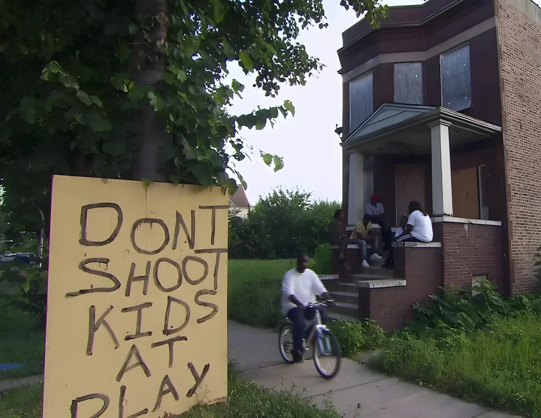Photo of a handwritten plywood sign leaning against a tree in an urban residential neighbourhood. Sign reads in large hand-painted letters: "DON'T SHOOT KIDS AT PLAY". Behind it, a child rides a bicycle on the sidewalk past a brick rowhouse where several people sit on the front steps