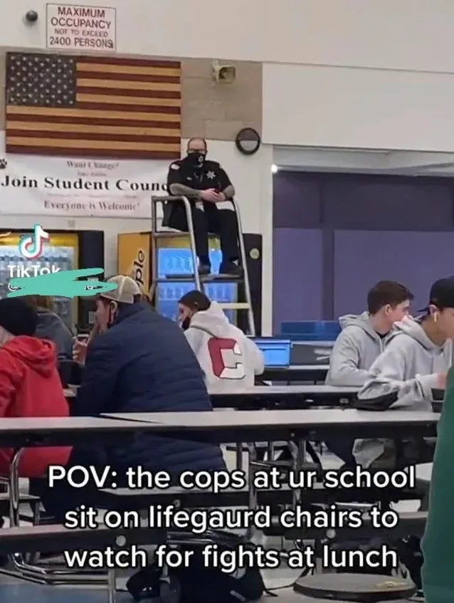 TikTok screenshot (username redacted) from inside a school cafeteria. A police officer in dark uniform sits elevated on a lifeguard-style high chair, overlooking students eating lunch. American flag artwork on the wall behind. "Maximum Occupancy Not To Exceed 2400 Persons" sign visible. "Join Student Council - Everyone is Welcome" banner. Caption: "POV: the cops at ur school sit on lifeguard chairs to watch for fights at lunch"