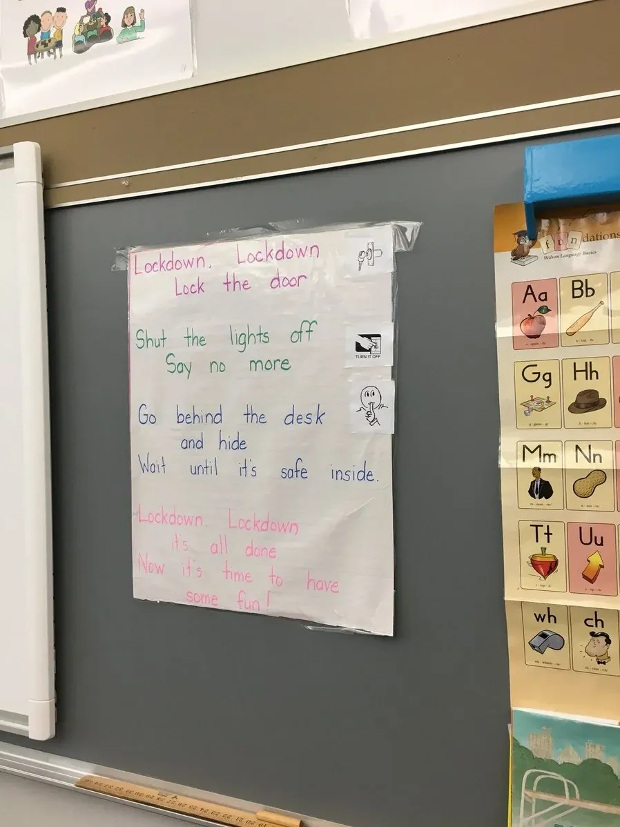 Photo of a handwritten poster taped to a classroom wall next to an alphabet chart. The poster is a lockdown drill poem written in coloured marker: "Lockdown, Lockdown / Lock the door / Shut the lights off / Say no more / Go behind the desk / and hide / Wait until it's safe inside / Lockdown, Lockdown / it's all done / Now it's time to have / some fun!"