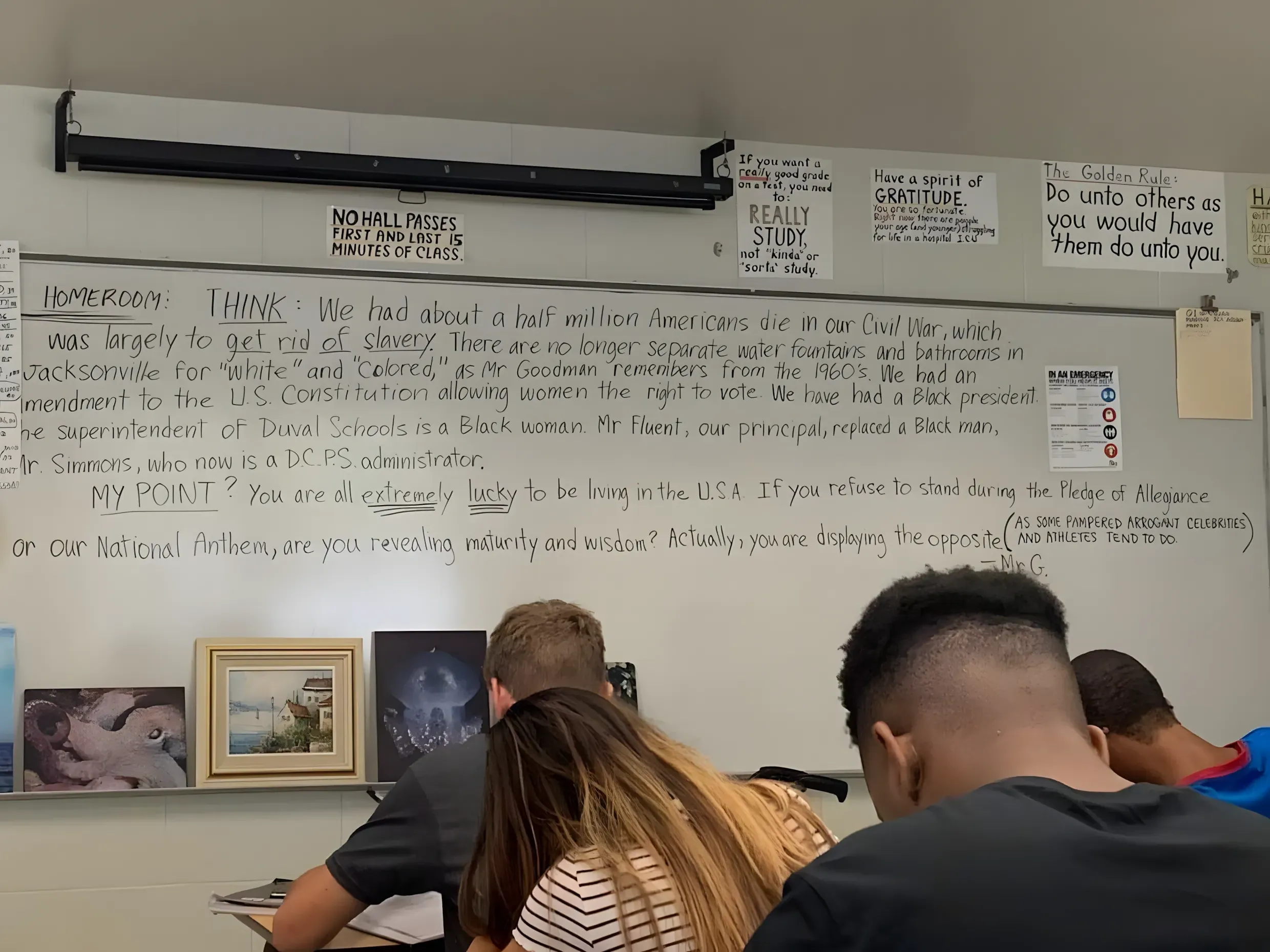 Photo of a classroom whiteboard with teacher's handwritten message in cursive. Text reads: "HOMEROOM: THINK: We had about a half million Americans die in our Civil War, which was largely to get rid of slavery. There are no longer separate water fountains and bathrooms in Jacksonville for 'White' and 'Colored,' as Mr Goodman remembers from the 1960's. We had an amendment to the U.S. Constitution allowing women the right to vote. We have had a Black president. The superintendent of Duval Schools is a Black woman. Mr Fluent, our principal, replaced a Black man, Mr Simmons, who now is a DCPS administrator. MY POINT? You are all extremely lucky to be living in the U.S.A. If you refuse to stand during the Pledge of Allegiance or our National Anthem, are you revealing maturity and wisdom? Actually, you are displaying the opposite." Additional note about pampered arrogant celebrities and athletes. Motivational signs and framed artwork visible above the whiteboard