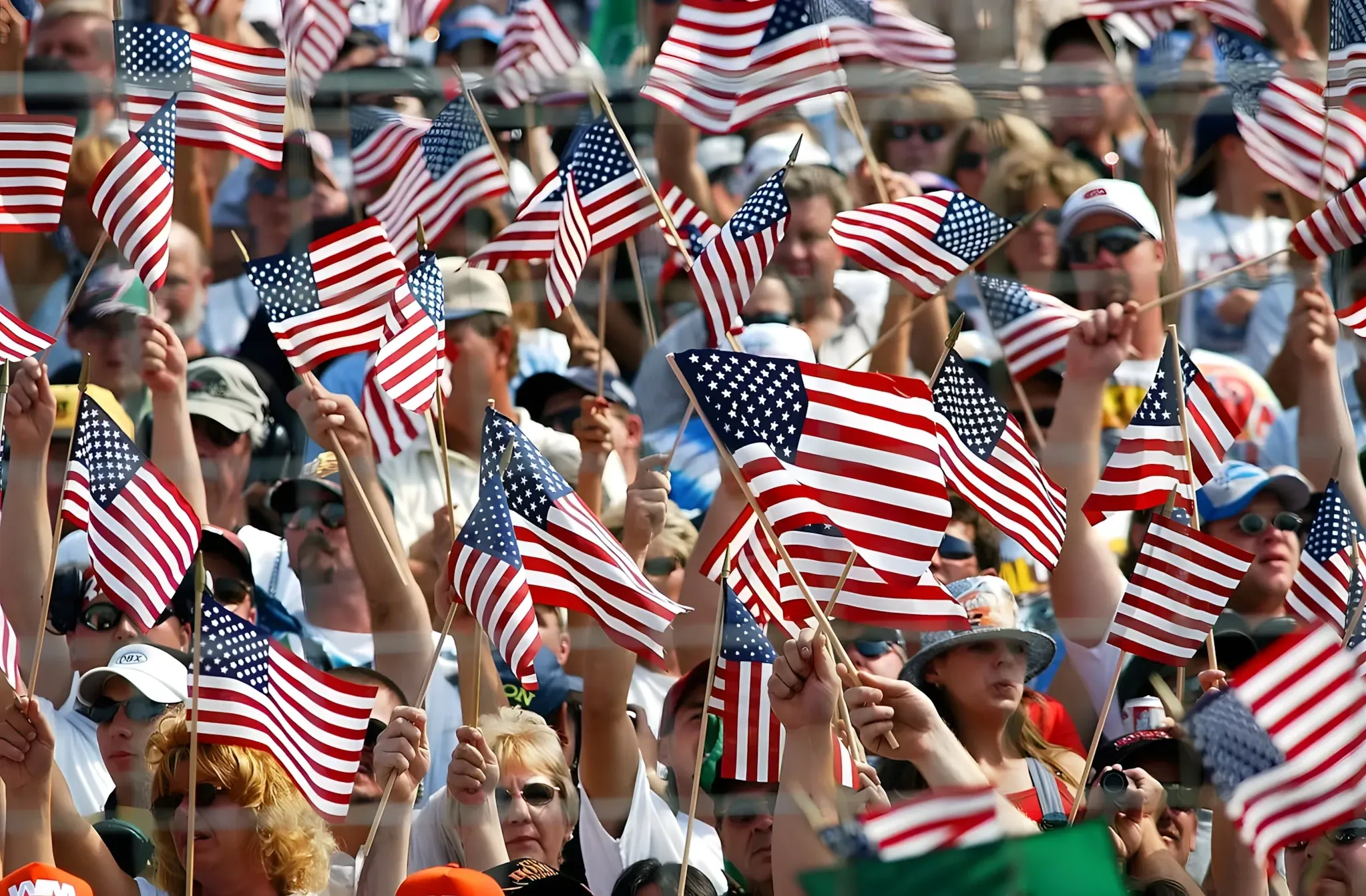 Photo of a large crowd at an outdoor event/stadium, with dozens of people waving small American flags. The crowd fills the entire frame, all holding flags in the air