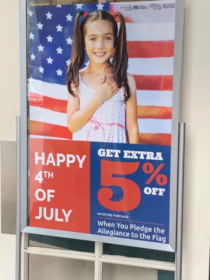 Photo of a store display sign. A young girl with pigtails stands in front of an American flag, hand on heart. Store logos: Perfume 4U, Perfume Europa, Gifts by Perfume 4U, Melhores Perfumes. Text: "HAPPY 4TH OF JULY" and "GET EXTRA 5% OFF ON ENTIRE PURCHASE When You Pledge the Allegiance to the Flag"
