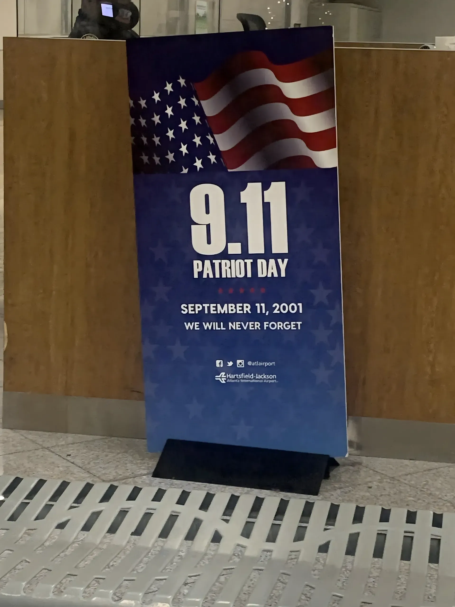 Photo of a tall standing banner/sign at Hartsfield-Jackson Atlanta International Airport. Blue gradient background with waving American flag at top. Text: "9.11 PATRIOT DAY" with five red stars. "SEPTEMBER 11, 2001 WE WILL NEVER FORGET". Social media icons and @atlairport handle. Hartsfield-Jackson logo at bottom. Sign on tiled floor near wooden-panelled walls