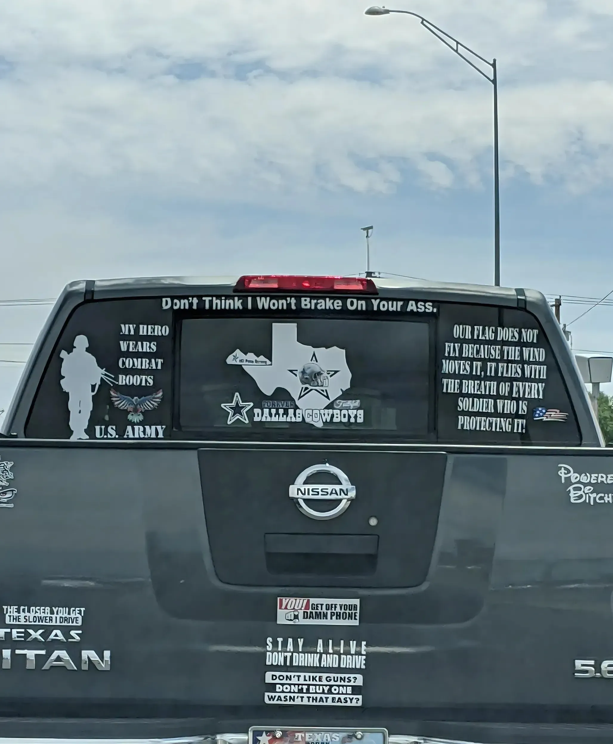 Photo of the rear of a black Nissan Titan pickup truck in Texas. Multiple patriotic and other stickers/decals. Top: "Don't Think I Won't Brake On Your Ass." Left: soldier silhouette with "MY HERO WEARS COMBAT BOOTS" and "U.S. ARMY". Centre: Dallas Cowboys logo inside Texas state outline. Right: "OUR FLAG DOES NOT FLY BECAUSE THE WIND MOVES IT, IT FLIES WITH THE BREATH OF EVERY SOLDIER WHO IS PROTECTING IT!" Bottom stickers: "THE CLOSER YOU GET THE SLOWER I DRIVE", "TEXAS TITAN", "YOU! GET OFF YOUR DAMN PHONE", "STAY ALIVE DON'T DRINK AND DRIVE", "DON'T LIKE GUNS? DON'T BUY ONE WASN'T THAT EASY?", "Powered by Bitch" partially visible