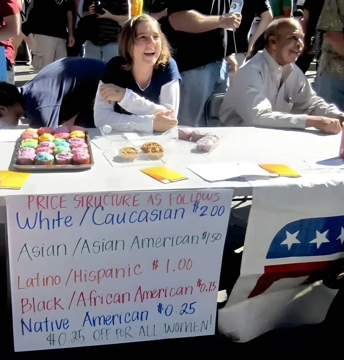 Photo of a bake sale at an outdoor event. A woman sits behind a table with colourful cupcakes. A handwritten sign reads: "PRICE STRUCTURE AS FOLLOWS White/Caucasian $2.00, Asian/Asian American $1.50, Latino/Hispanic $1.00, Black/African American $0.75, Native American $0.25, $0.25 OFF FOR ALL WOMEN!" Republican party logo visible on the right side of the table