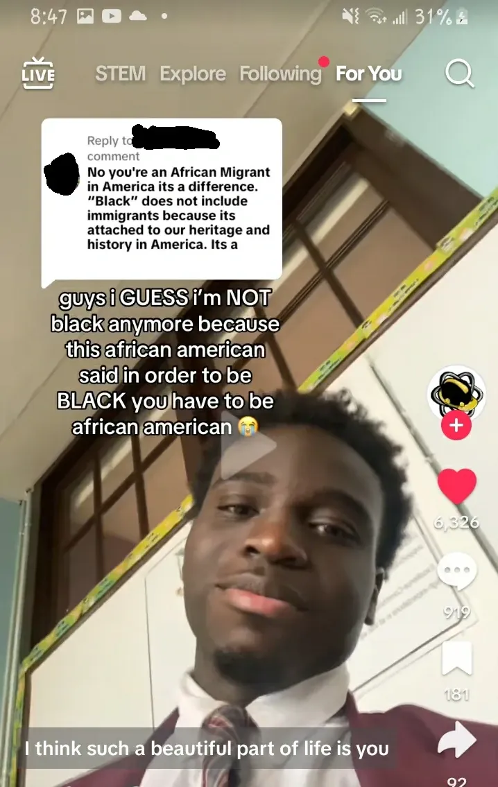 TikTok screenshot on mobile. For You page. Video of a young black man in a shirt and tie with text overlay: "guys i GUESS i'm NOT black anymore because this african american said in order to be BLACK you have to be african american" with crying emoji. Overlaid comment: "No you're an African Migrant in America its a difference. 'Black' does not include immigrants because its attached to our heritage and history in America. Its a" (cut off).