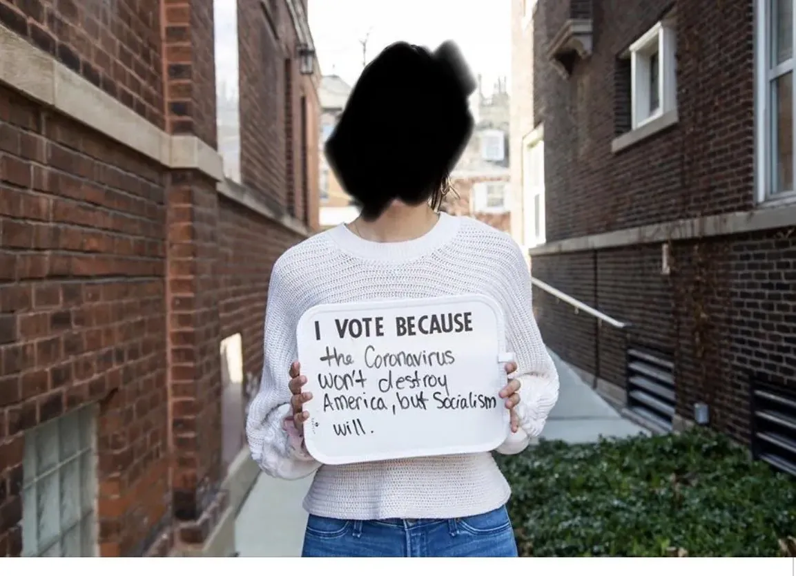 Photo of a person (face redacted) standing in an alley between brick buildings, holding a small whiteboard reading "I VOTE BECAUSE the Communists were planning America into Socialism went..." (partially illegible handwritten text). Person wearing a white sweater and blue jeans