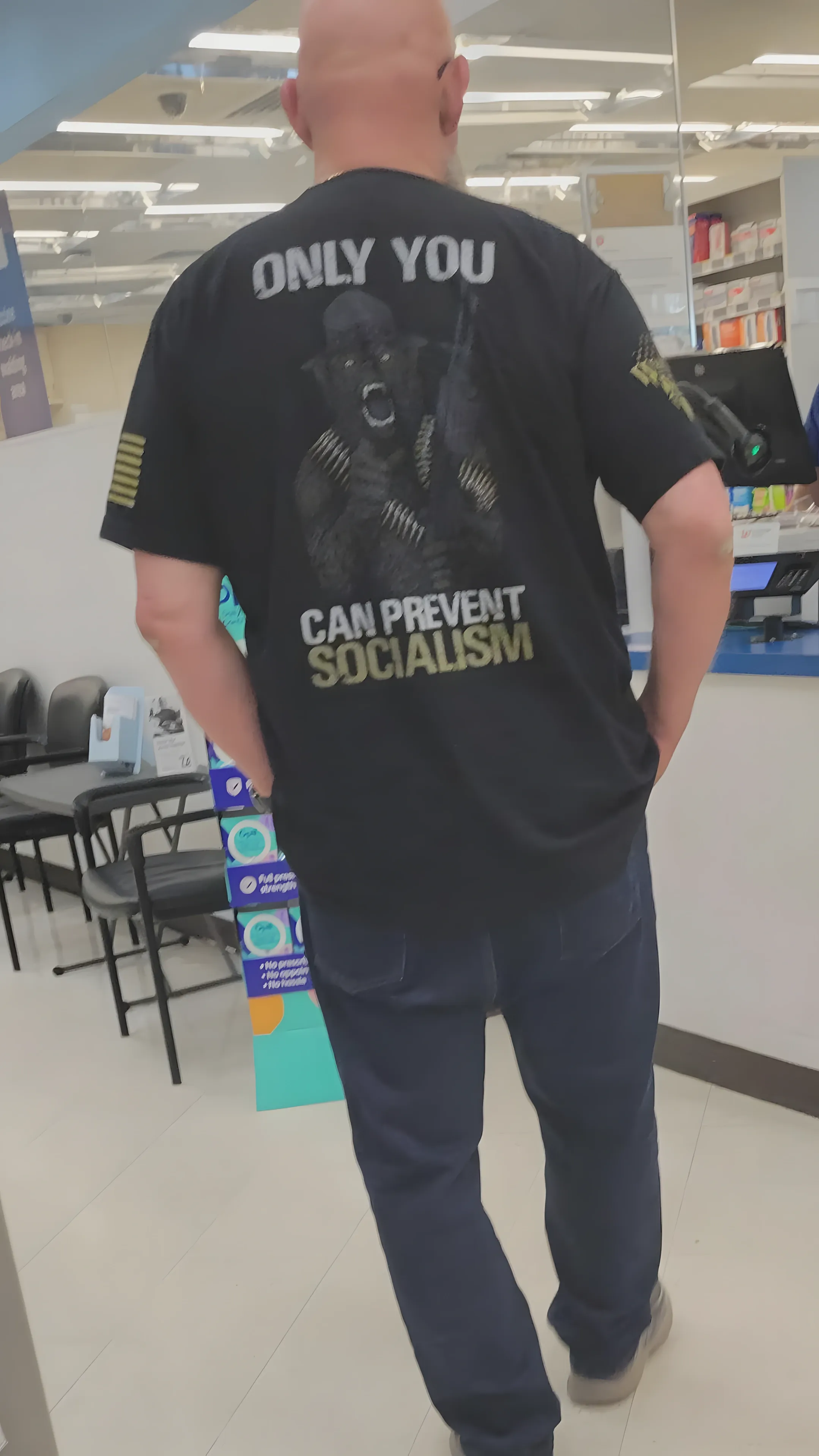 Photo of a bald man standing in a pharmacy/store, photographed from behind. He's wearing a black t-shirt with an image of a soldier holding a rifle and text reading "ONLY YOU CAN PREVENT SOCIALISM". American flag detail on the sleeve. Blue jeans and grey shoes