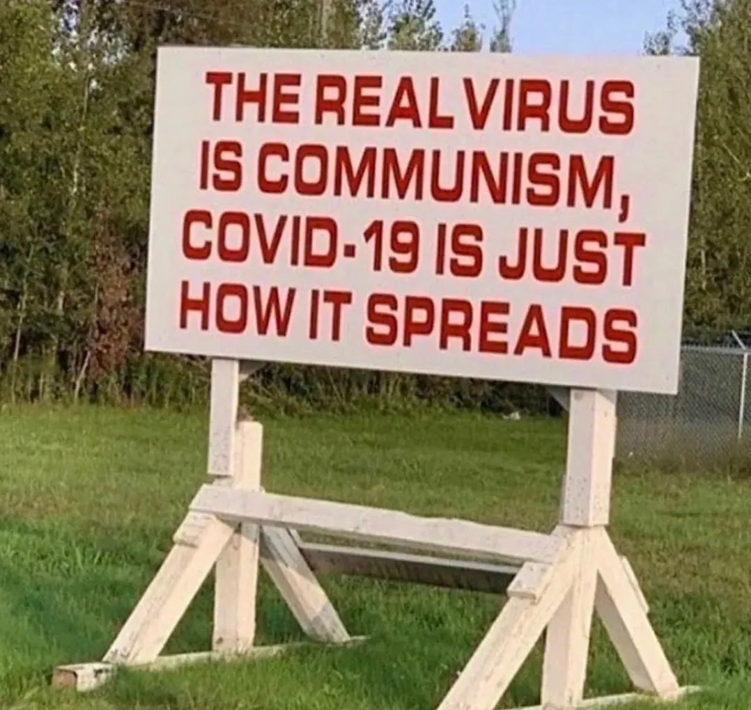 Photo of a white wooden roadside sign on green grass. Red text: "THE REAL VIRUS IS COMMUNISM, COVID-19 IS JUST HOW IT SPREADS"
