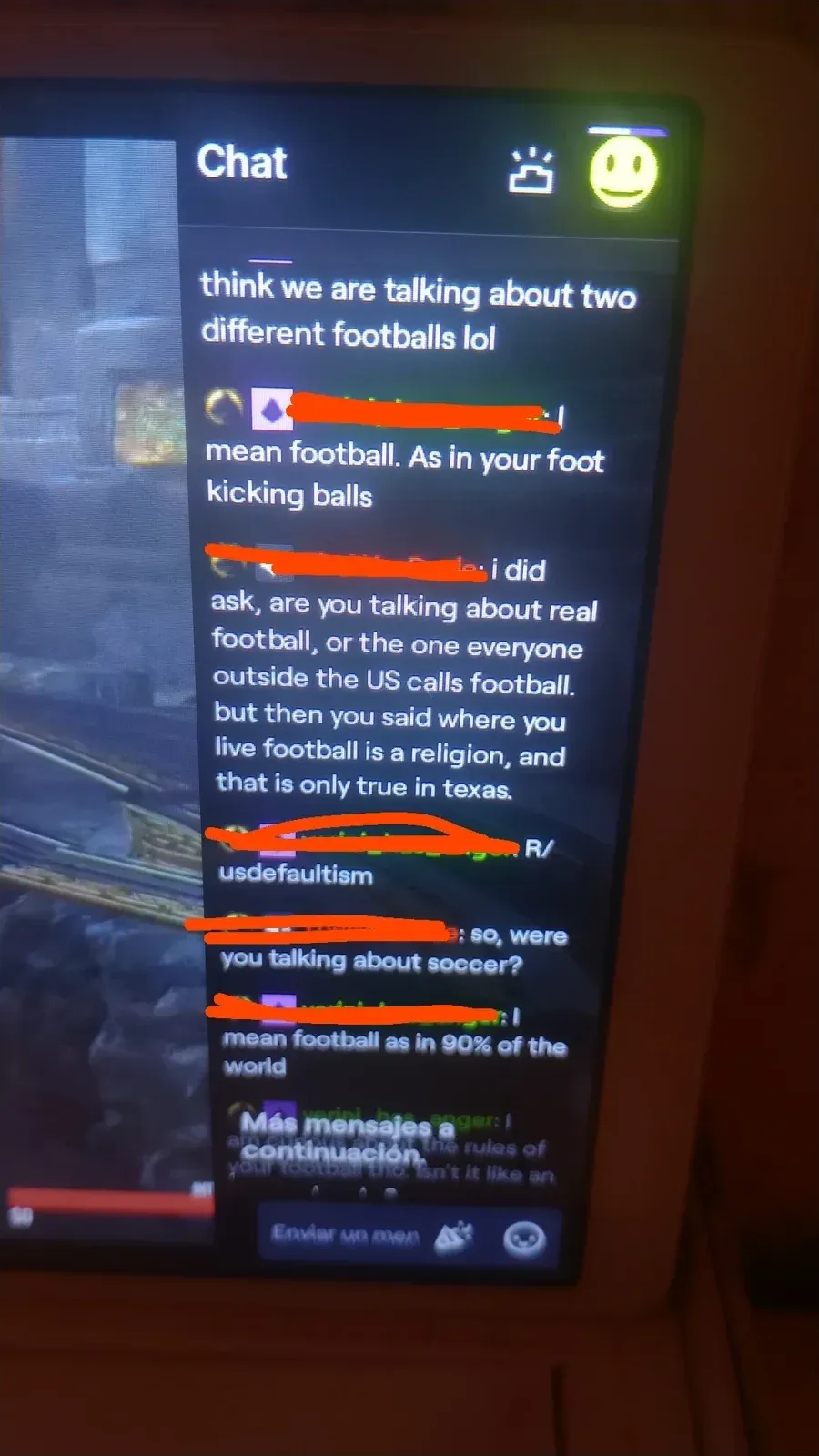 Photo of a TV/monitor screen showing a gaming chat. Conversation between users (username redacted): "think we are talking about two different footballs lol". Green: "I mean football. As in your foot kicking balls". Red: "I did ask, are you talking about real football, or the one everyone outside the US calls football. But then you said where you live football is a religion, and that is only true in texas." Red links: "r/usdefaultism". Green: "so, were you talking about soccer?" Another user: "mean football as in 90% of the world". Spanish text visible at bottom