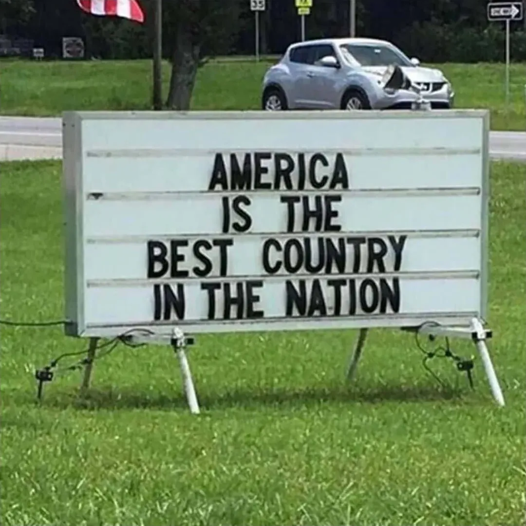 Photo of a roadside letter board sign on grass verge with an American flag on top. The sign reads: "AMERICA IS THE BEST COUNTRY IN THE NATION". A white SUV is parked behind it