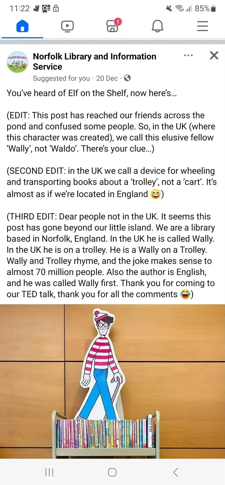 Facebook mobile screenshot. Norfolk Library and Information Service post (Suggested for you, 20 Dec) with photo of a cardboard cutout of Wally (Where's Wally character in red and white striped outfit). Post: "You've heard of Elf on the Shelf, now here's..." Three edits: "(EDIT: This post has found our friends across the pond and confused some people. So, in the UK (where this character was created), we call this elusive fellow 'Wally', not 'Waldo'. There's your clue.)" "(SECOND EDIT: in the UK we call a device for wheeling and transporting books about a 'trolley', not a 'cart'. It's almost as if we're located in England)" "(THIRD EDIT: Dear people not in the UK. It seems this post has gone beyond our little island. We are a library based in Norfolk, England. In the UK he is called Wally. In the UK he is on a trolley. Wally and Trolley rhyme, and the joke makes sense to almost 70 million people. Also the author is English, and he was called Wally first. Thank you for coming to our TED talk, thank you for all the comments)"