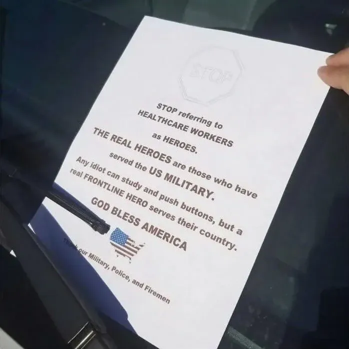 Printed flyer held up against a car window. Text reads: "STOP referring to HEALTHCARE WORKERS as HEROES. THE REAL HEROES are those who have served the US MILITARY. Any idiot can study and push buttons, but a real FRONTLINE HERO serves their country. GOD BLESS AMERICA" with a US flag map graphic and "Thank our Military, Police, and Firemen" at the bottom.