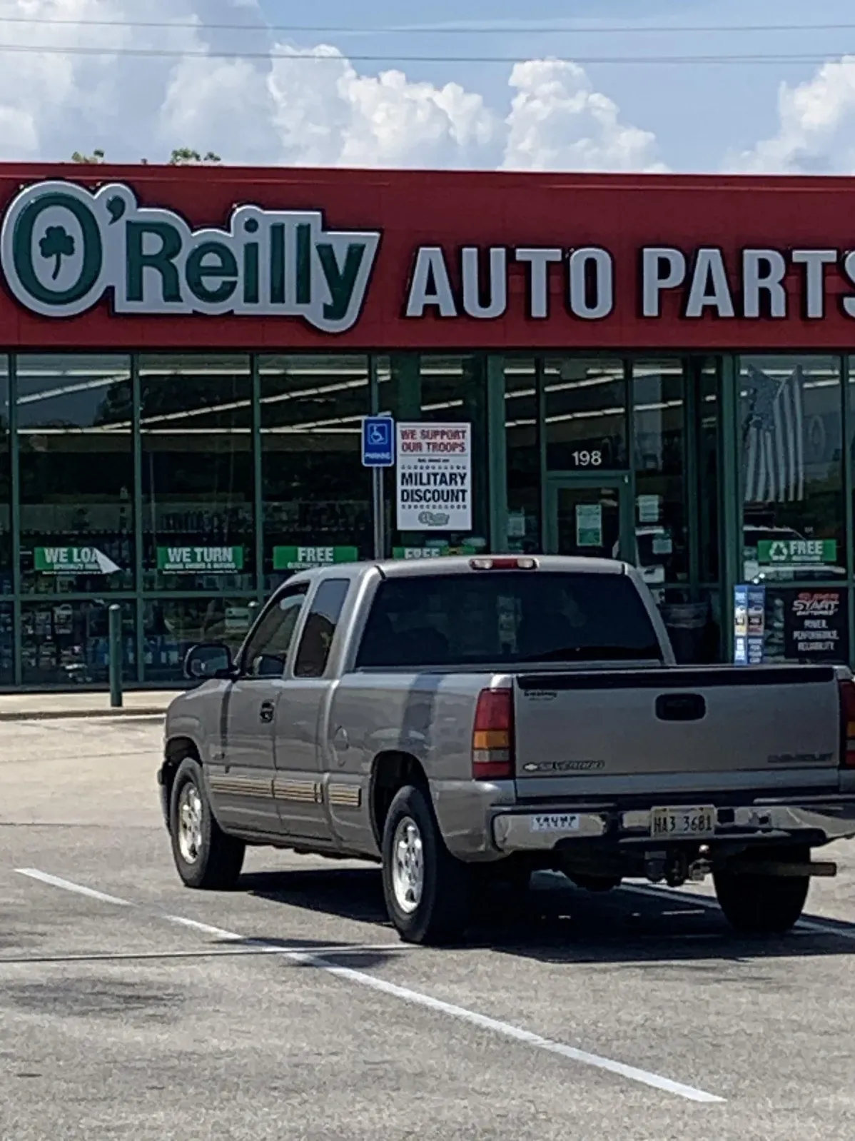 Photo of an O'Reilly Auto Parts store (red storefront with green shamrock logo, store #198) with a silver Chevrolet Silverado pickup truck parked in front. Signs in the window include "WE SUPPORT OUR TROOPS" and "MILITARY DISCOUNT".
