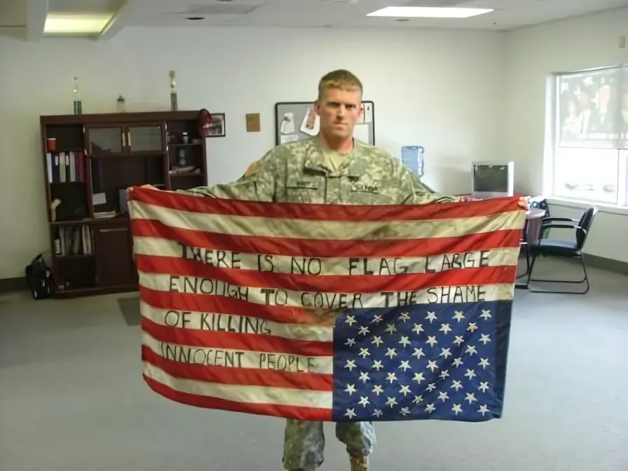 Photo of a US Army soldier in ACU camouflage uniform standing in an office or break room, holding an upside-down American flag with handwritten text on the stripes: "THERE IS NO FLAG LARGE ENOUGH TO COVER THE SHAME OF KILLING INNOCENT PEOPLE". The soldier stares directly at the camera with a serious expression.