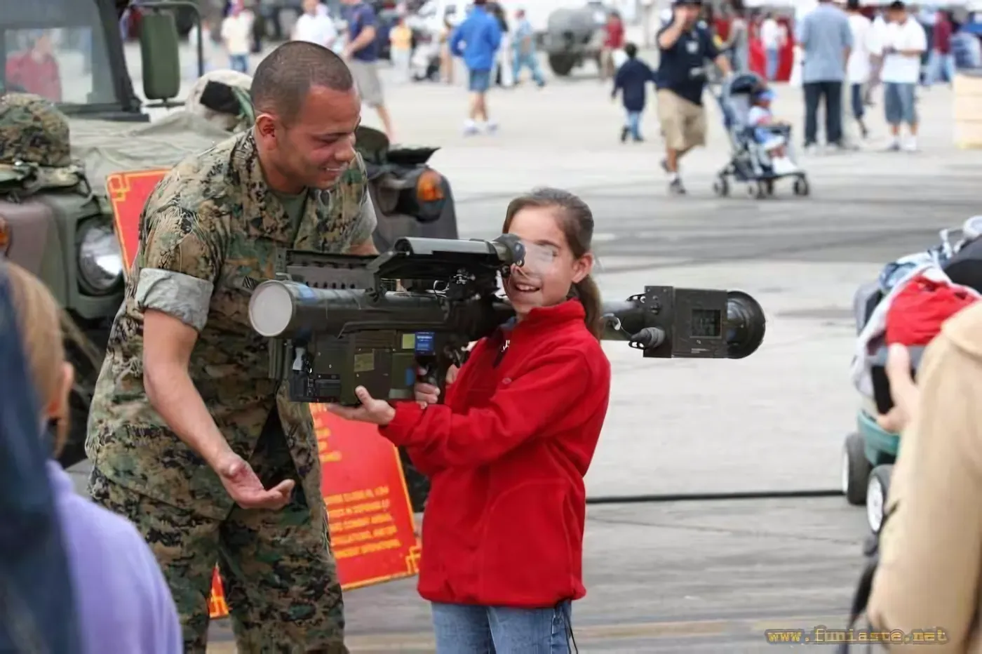 Photo from a military open day or airshow (watermark: www.funiaste.net). A young girl in a red jacket and safety glasses holds a large shoulder-fired military weapon (appears to be a Stinger missile launcher) while a smiling US Marine in MARPAT camouflage stands beside her. Crowd of civilians and military vehicles in the background.