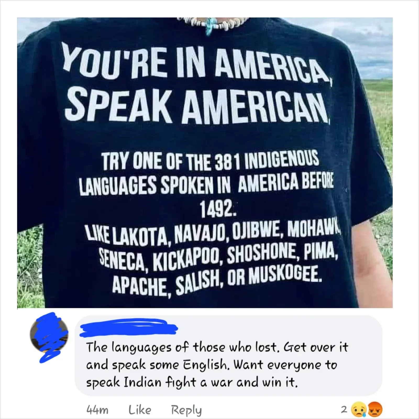 Facebook post. Photo of someone wearing a black t-shirt outdoors: "YOU'RE IN AMERICA, SPEAK AMERICAN. TRY ONE OF THE 381 INDIGENOUS LANGUAGES SPOKEN IN AMERICA BEFORE 1492. LIKE LAKOTA, NAVAJO, OJIBWE, MOHAWK, SENECA, KICKAPOO, SHOSHONE, PIMA, APACHE, SALISH, OR MUSKOGEE." Comment below (44m, 2 angry/sad reactions): "The languages of those who lost. Get over it and speak some English. Want everyone to speak Indian fight a war and win it."