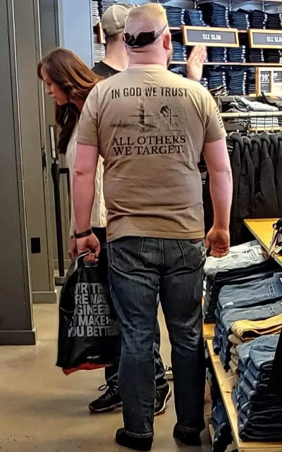 Photo taken in a clothing store (shelves of jeans labelled "5.11 SLIM"). A heavyset man seen from behind wearing a tan t-shirt with a rifle scope crosshair graphic and text: "IN GOD WE TRUST. ALL OTHERS WE TARGET." He's wearing jeans, a backwards cap, and sunglasses on his head. A woman stands beside him. He's carrying a black shopping bag with partially visible text "...IRTH ...RE WA... ...GINEER... ...MAKE... ...OU BETTE..."