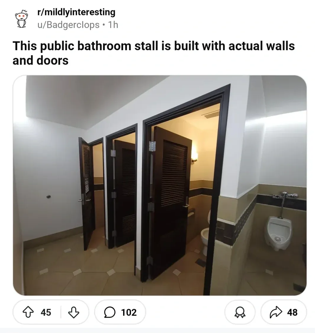 Reddit post from r/mildlyinteresting by u/Badgerclops. Title: This public bathroom stall is built with actual walls and doors. Photo shows elegant bathroom stalls with full floor-to-ceiling dark wood louvered doors and solid walls, built like actual rooms, with a urinal visible to the right.