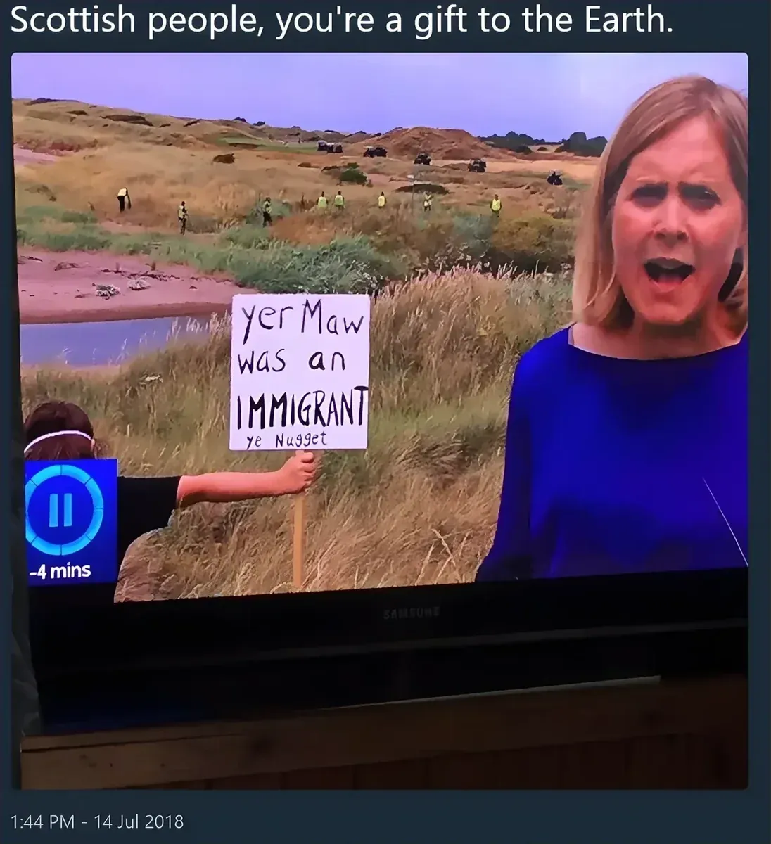 Tweet - Scottish people, you're a gift to the Earth. Photo of a TV news broadcast from Trump's Turnberry golf course showing a protester holding a handwritten sign reading "yer Maw was an IMMIGRANT ye Nugget" with a female reporter in the foreground. 14 Jul 2018