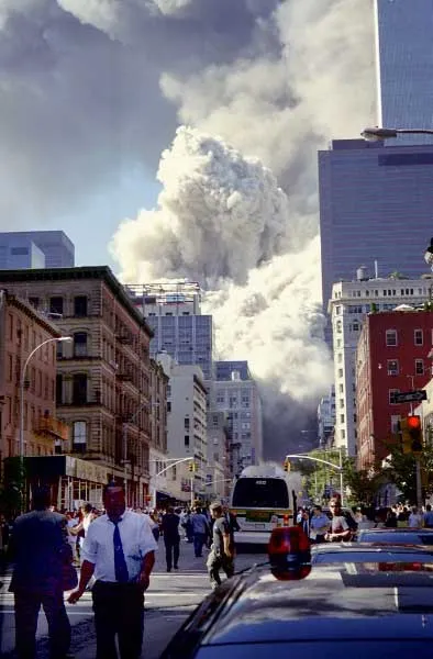 Street-level view looking down a Manhattan street toward the WTC collapse, massive dust cloud rising between buildings, pedestrians and vehicles on the street, man in white shirt walking in the foreground