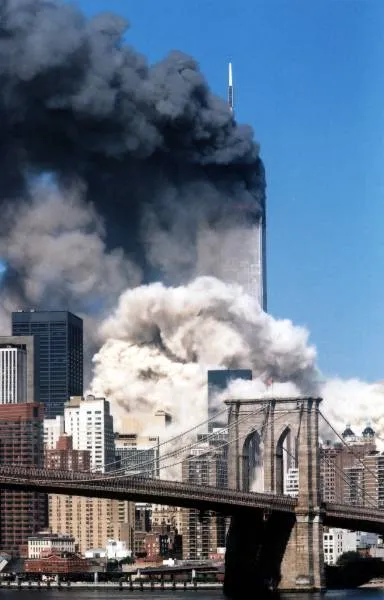 View of WTC towers with one still standing amid heavy smoke and dust, seen behind the Brooklyn Bridge towers and cables, blue sky above