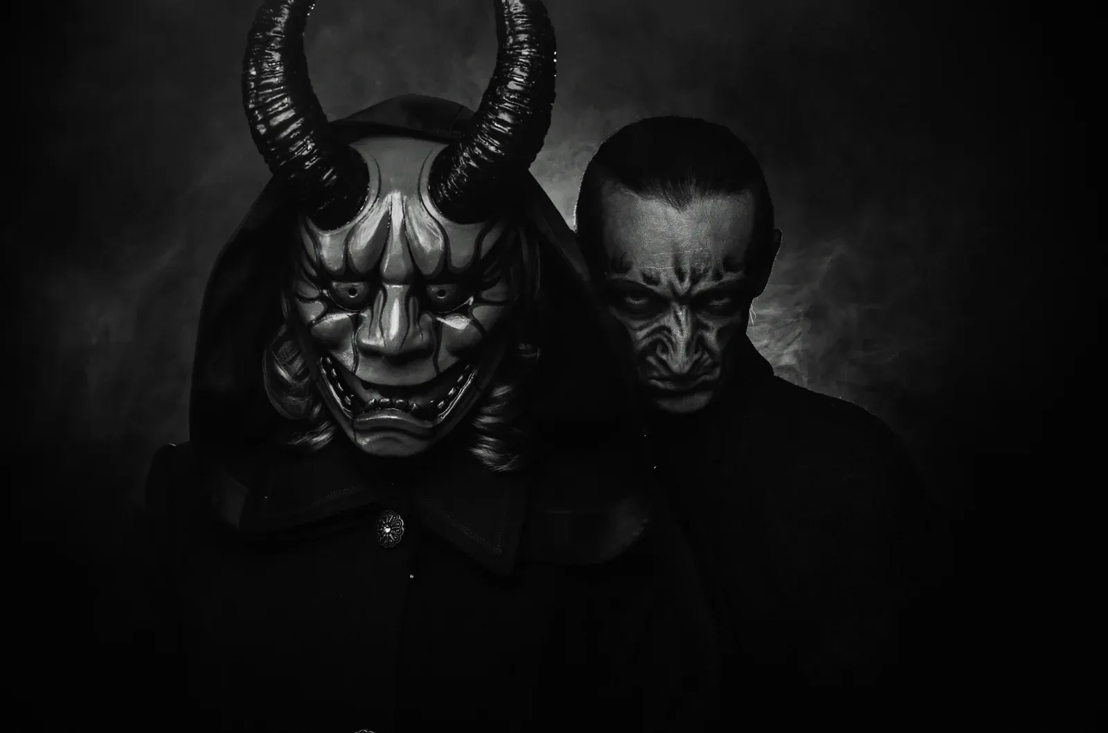 Two sinister figures in darkness, one wearing a horned Japanese oni mask and the other with demonic face paint, shrouded in smoke