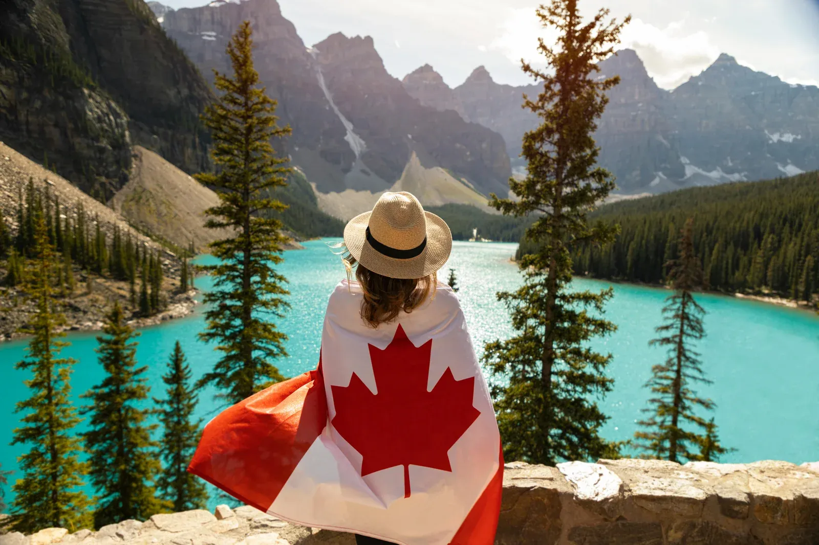 Woman draped in a Canadian flag looking out over the turquoise water of Moraine Lake with the Rocky Mountains behind her