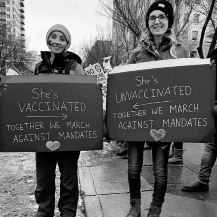 Black and white photo of two women at anti-mandate protest holding signs with arrows pointing at each other - She's VACCINATED Together We March Against Mandates and She's UNVACCINATED Together We March Against Mandates, with heart symbols