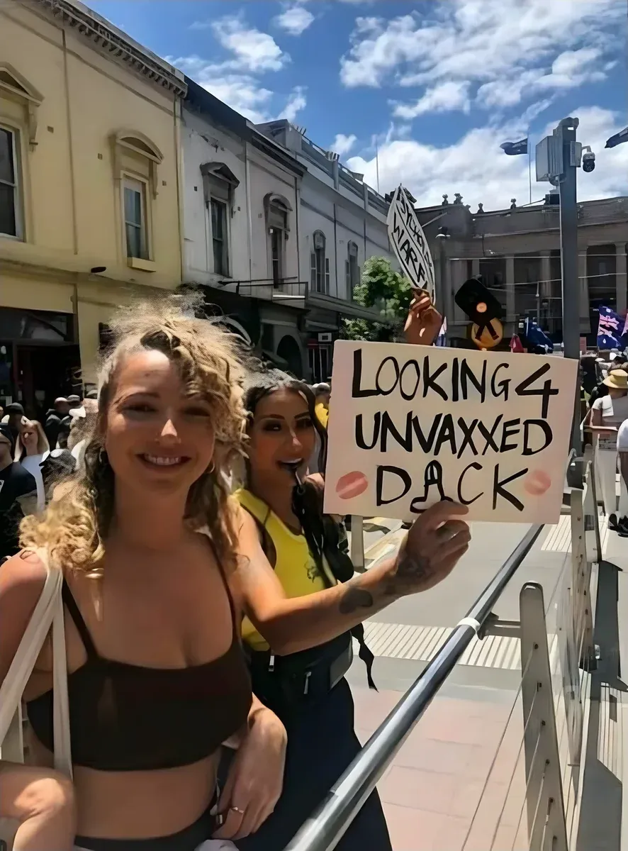 Photo from Australian anti-mandate protest - two young women smiling at camera, one holding handwritten sign reading LOOKING 4 UNVAXXED DACK (with kiss mark stickers). Crowd and Australian flags visible in background, sunny day in what appears to be a town centre